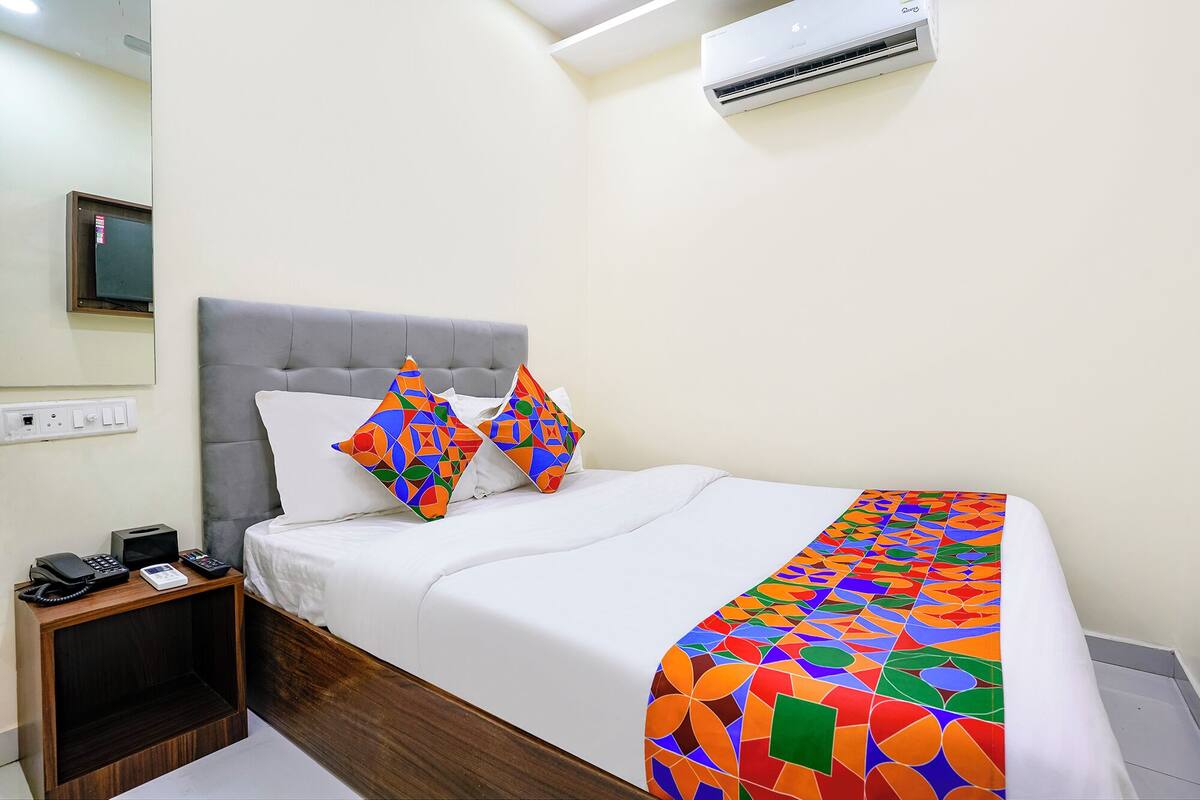 A neatly made bed with white linens, two colorful geometric-patterned pillows, a wooden bedside table with a phone and a small electronic device, and a wall-mounted air conditioner.