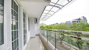 A balcony with a glass railing, a white exterior wall, and a view of a cityscape with buildings and trees. - Fabhotel Trident (New Delhi)