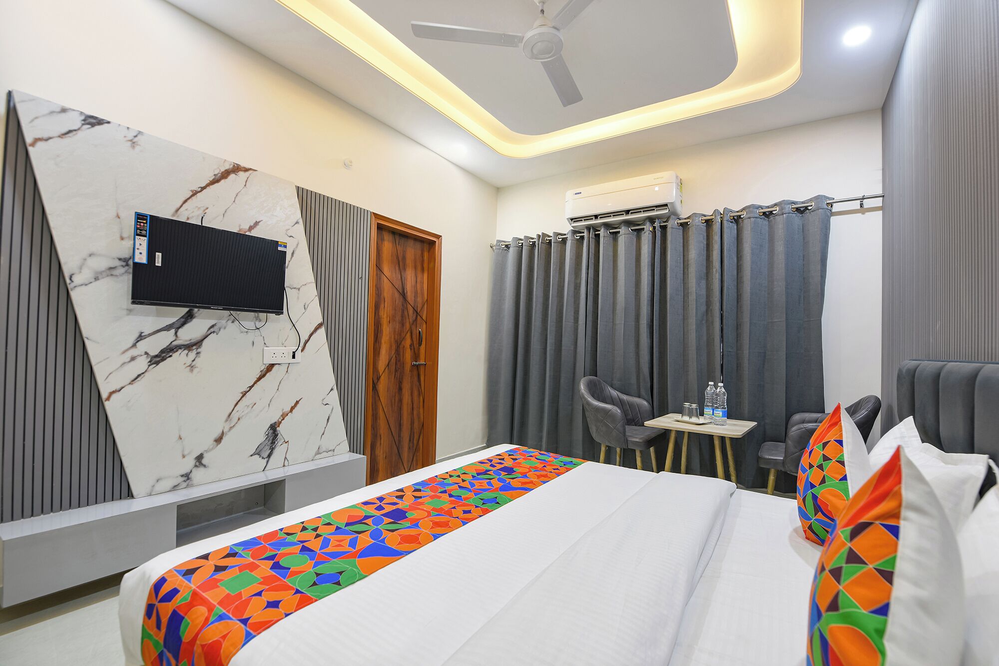 A hotel room with a bed, a TV mounted on a marble wall, a ceiling fan, an air conditioning unit, a small table with chairs, and a window with curtains.