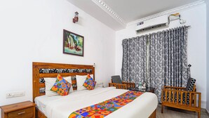 Een slaapkamer met een houten bed, kleurrijke kussens, een beddengoed met een patroon, een nachtkastje, een stoel, een raam met gordijnen met dessin en een airconditioning.
