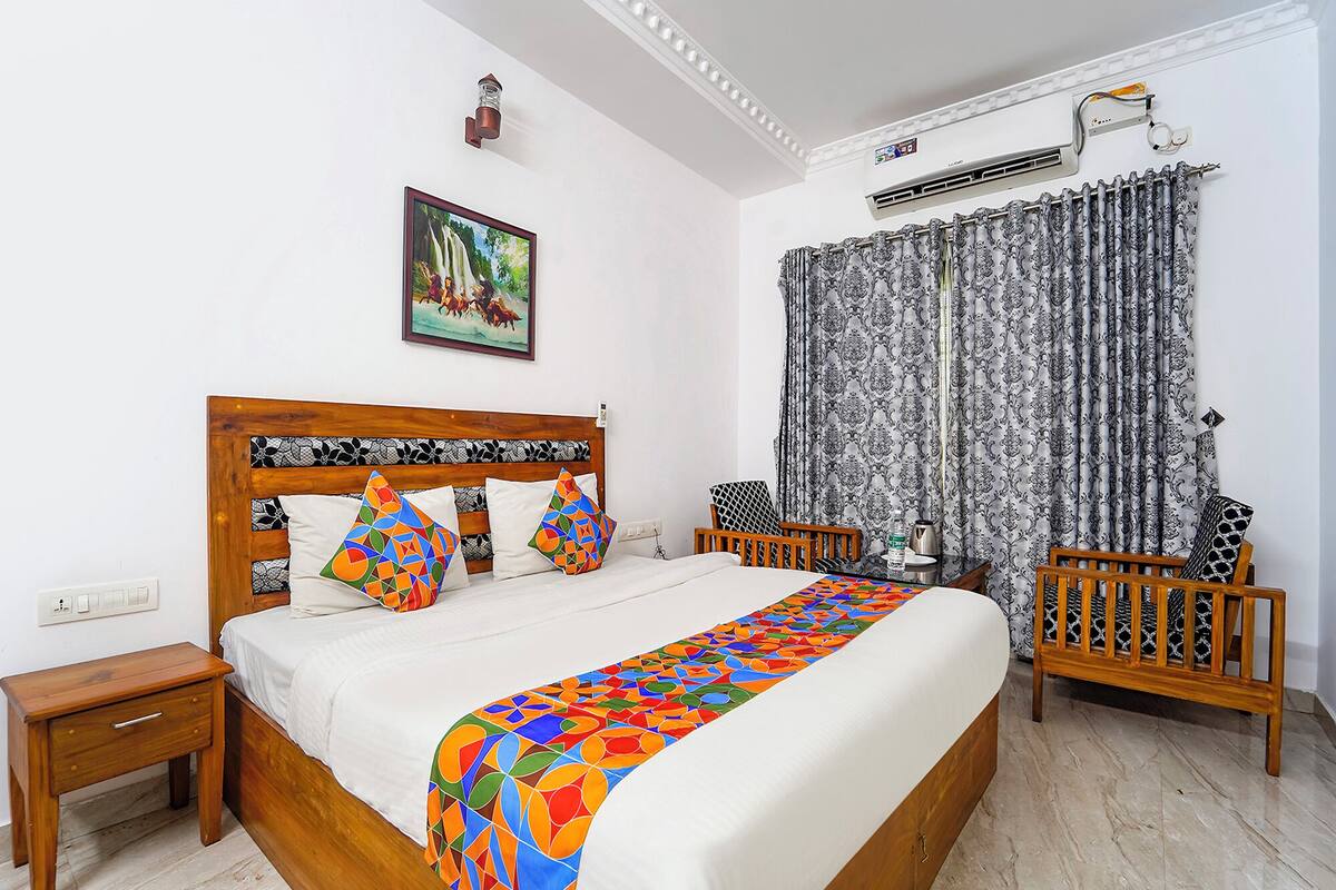 A bedroom with a wooden bed, colorful pillows, a patterned bedspread, a nightstand, a chair, a window with patterned curtains, and an air conditioning unit.