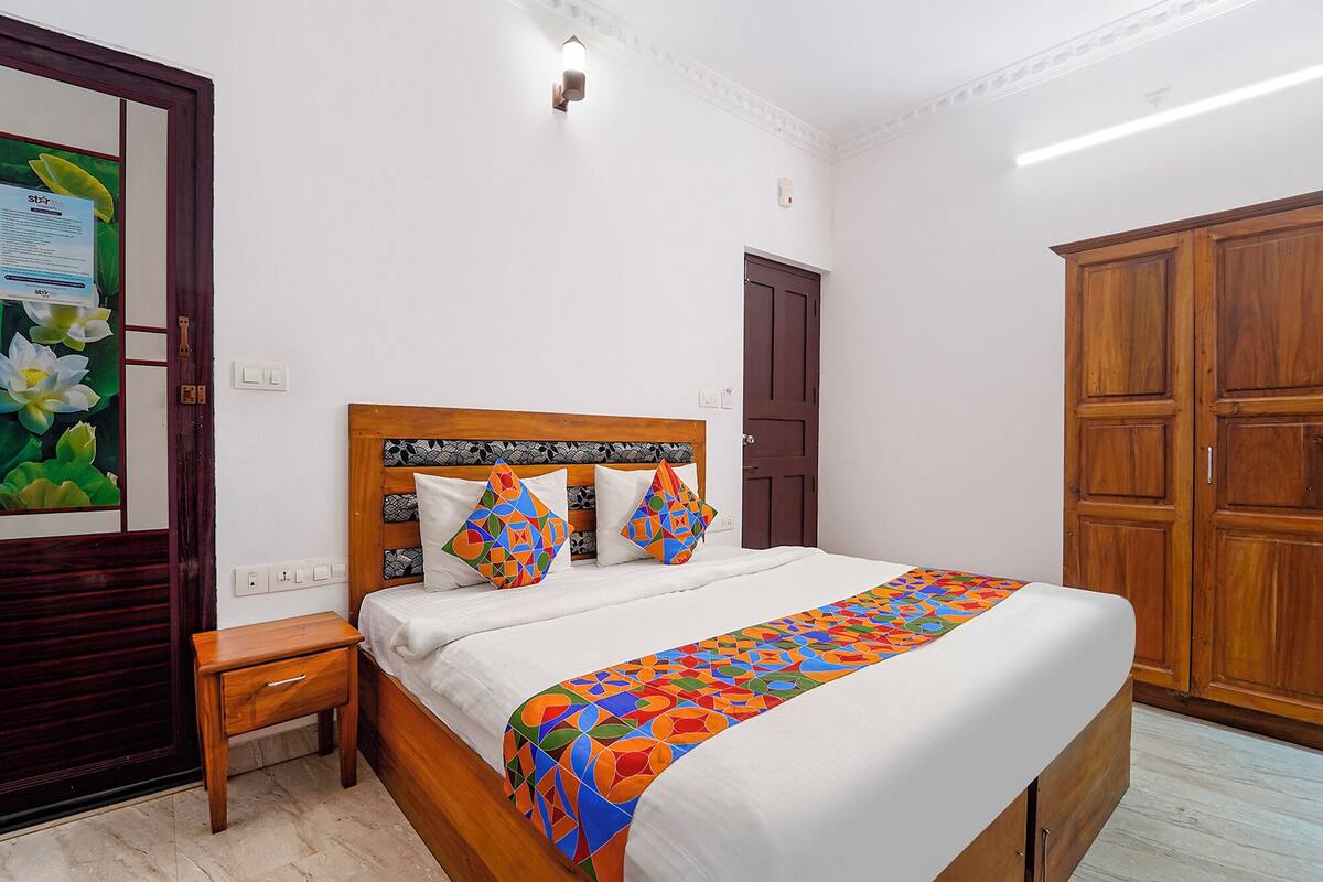 A bedroom with a wooden bed, white bedding, and colorful pillows. There is a wooden nightstand, a door, and a wardrobe.