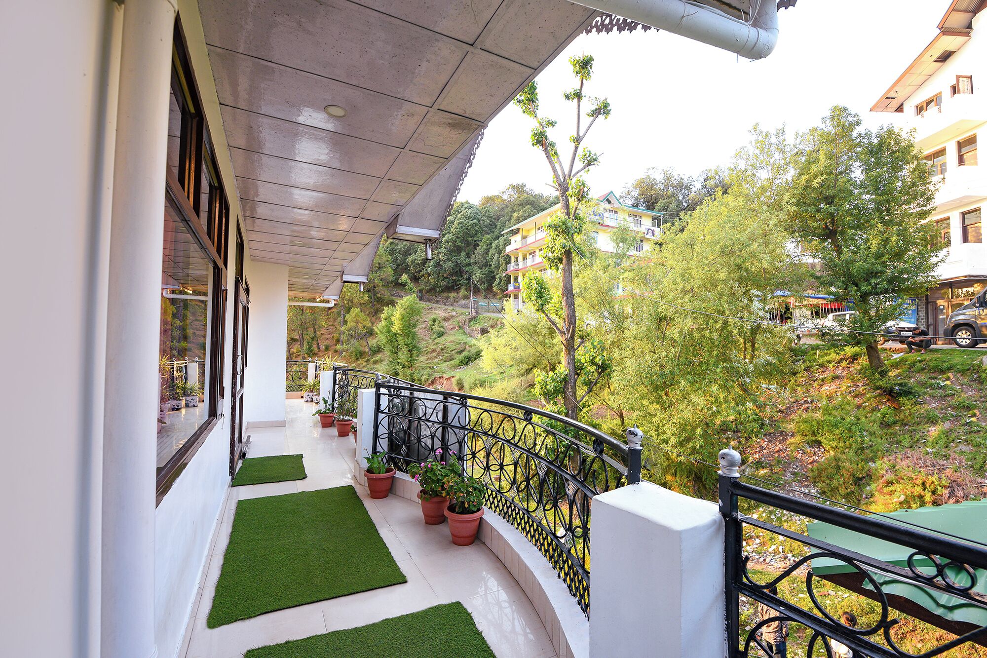 A balcony with artificial grass, potted plants, and a metal railing overlooking a hillside with trees and buildings.
