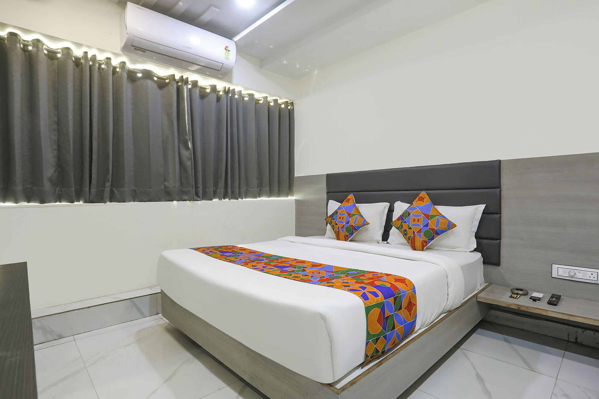 A modern hotel room with a large bed, grey curtains, an air conditioning unit, and a bedside table with a phone and remote controls.