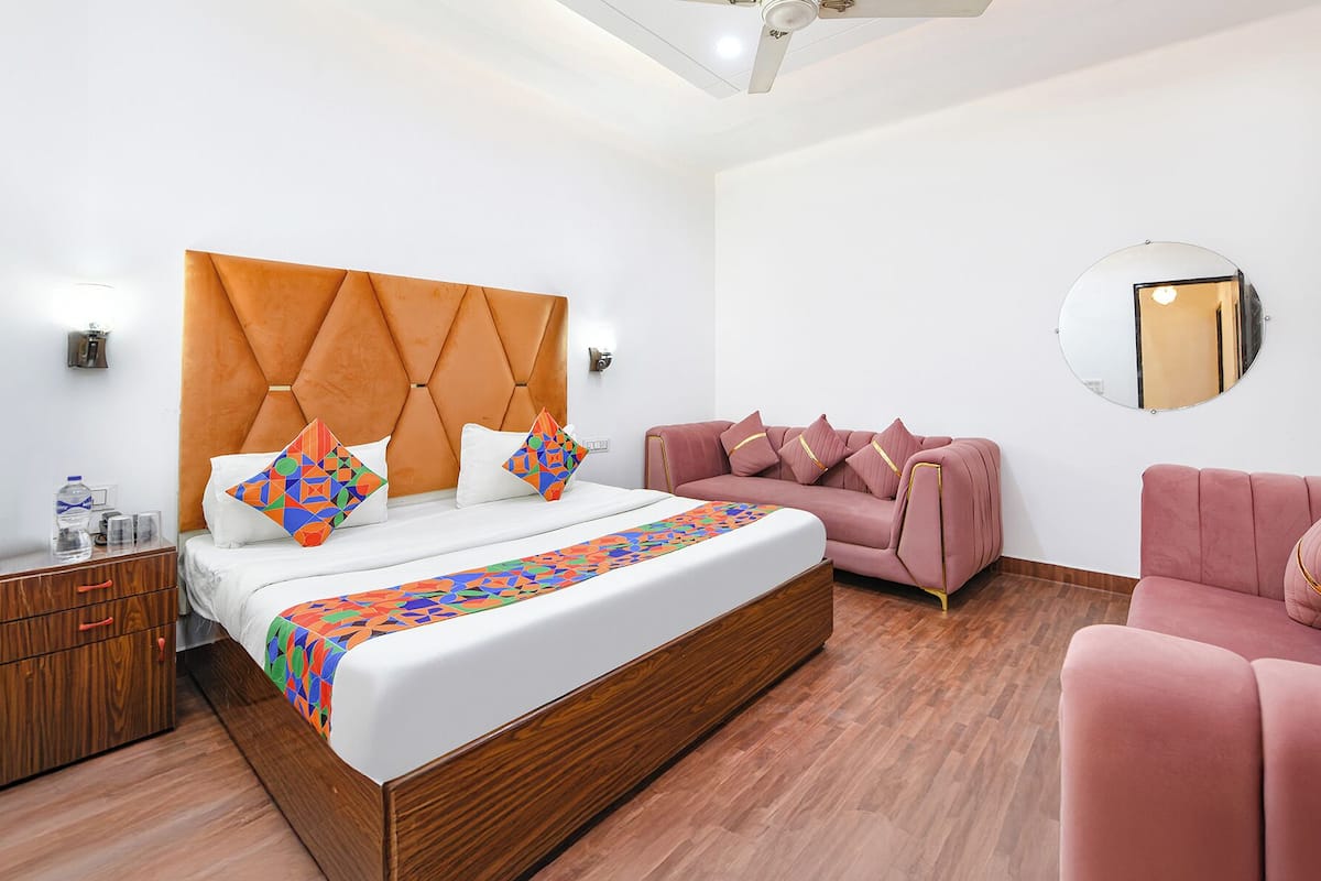 Moderni hotellihuone, jossa on suuri sänky, vaaleanpunainen sohva, puinen yöpöytä ja pyöreä peili seinällä.