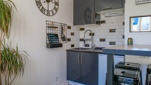 A modern kitchen with a black countertop, white tiled backsplash, and a black refrigerator. There is a black bar stool and a potted plant in the corner. - Charming Studio for 2/4 Guests – City Center (Les Sables-d´Olonne)
