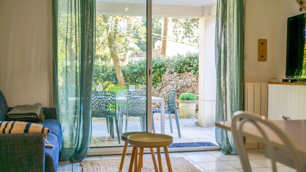 A living room with a sofa, a small stool, and a dining table with chairs. There is a sliding glass door leading to an outdoor area with a table and chairs.