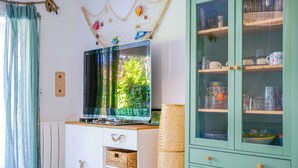 A room with a television on a wooden cabinet, a green cabinet with glass doors, and a window with a blue curtain.