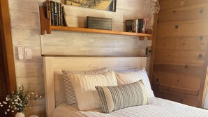 Una camera da letto con una testiera in legno, un letto con cuscini a righe e con texture, un comodino con un vaso e una sveglia, e un quadro sopra il letto.