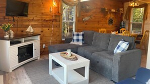 Un accogliente interno di una baita con un divano grigio, un tavolino bianco, una TV a parete e un camino.