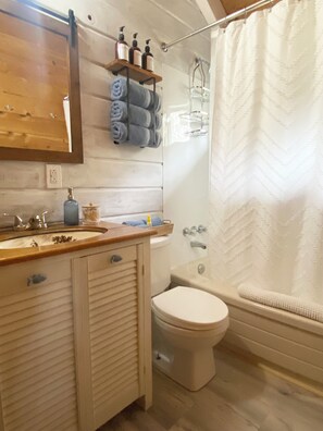 A bathroom with a wooden vanity, a toilet, a bathtub, and a shower curtain. - Pondside Haven (Tors Cove)