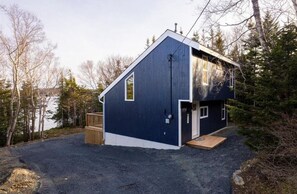 A two-story cabin with a dark blue exterior, a wooden deck, and a gravel driveway. - Pondside Haven (Tors Cove)