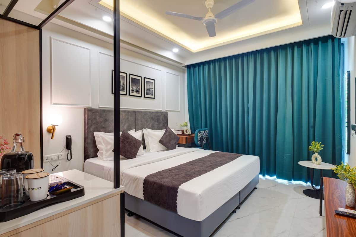 A modern hotel room with a large bed, a bedside table, a desk, a chair, a small table with a vase, and a ceiling fan.