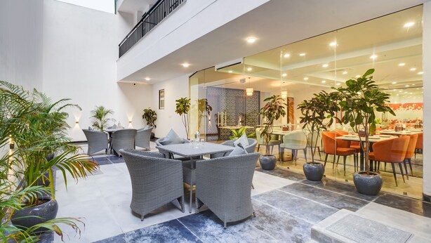 A modern indoor courtyard with wicker furniture, potted plants, and a glass wall leading to a dining area. - Saltstayz Premier - Arjun Marg (Gurugram)