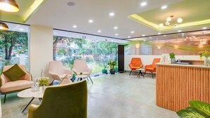 A modern reception area with a wooden reception desk, several seating options including a green chair, and large windows offering a view of the outdoors. - Saltstayz Premier - Arjun Marg (Gurugram)