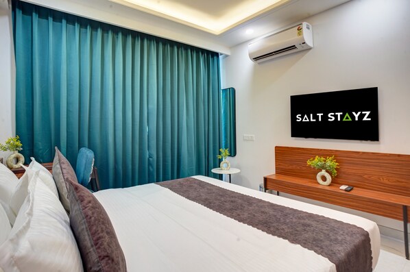 A modern hotel room with a bed, a TV mounted on the wall, a small table with a vase, and a bench with a plant. - Saltstayz Premier - Arjun Marg (Gurugram)