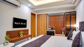 A modern hotel room with a large bed, a wooden bench, a small table with a plant, a TV mounted on the wall, and a wardrobe with a mirror.