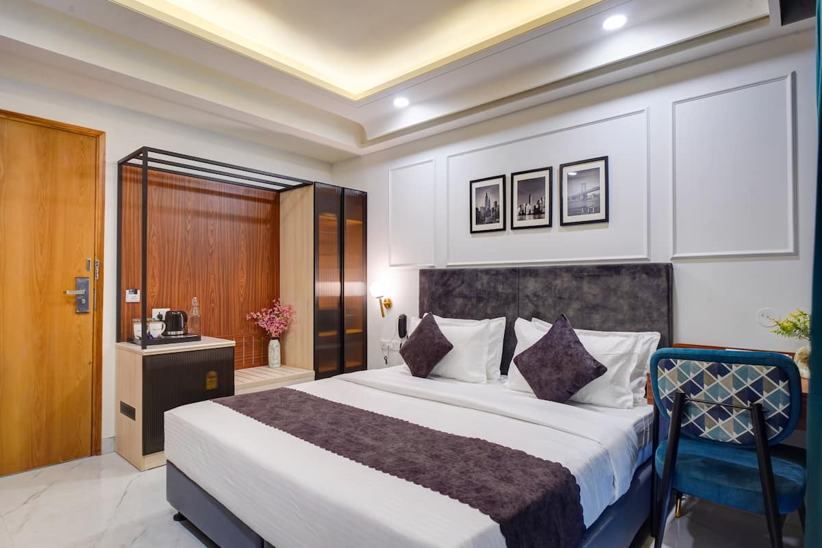 A modern hotel room with a large bed, a wooden wardrobe, a desk with a kettle, and a wall-mounted TV.