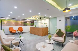 A modern reception area with a wooden reception desk, orange and white chairs, and a green wall with a plant. - Saltstayz Premier - Arjun Marg (Gurugram)