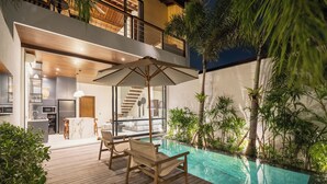 Ein moderner Poolbereich im Freien mit Holzterrasse, Liegestühlen und einer im Hintergrund erkennbaren, gläsernen Küche.