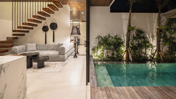 Ein modernes Interieur mit einer Holztreppe, einem grauen Sofa und einem Poolbereich mit tropischen Pflanzen.