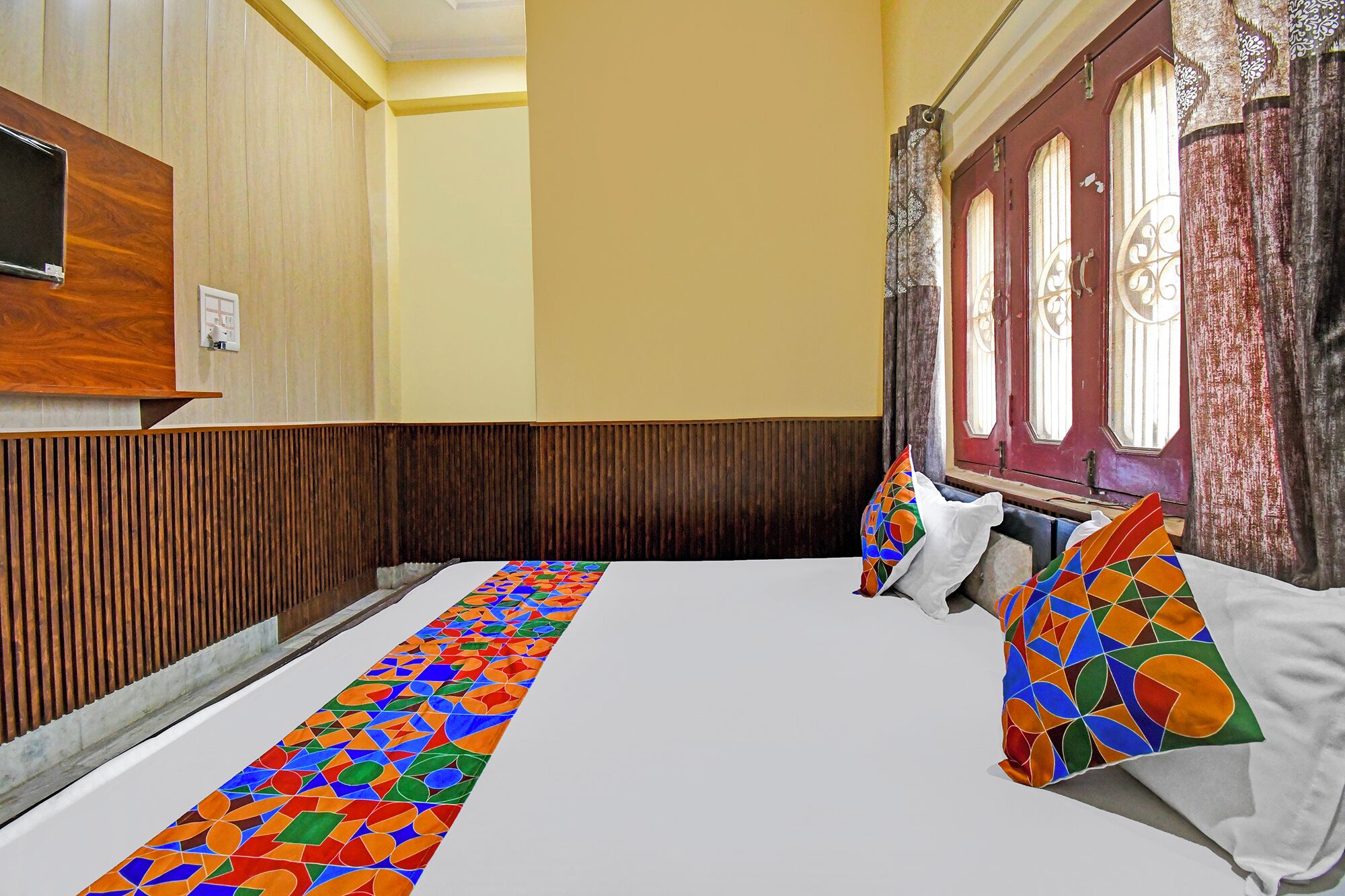 A hotel room with a wooden panel wall, a large bed with colorful pillows, and a window with curtains.