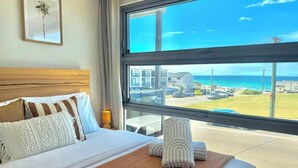 A hotel room with a large window, a bed with white linens, striped pillows, and a woven mat. - Merewether By The Beaches Newcastle, Free Parking (Merewether)