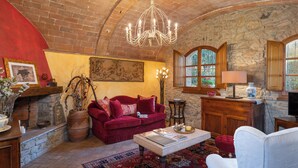A cozy living room with a red sofa, a white armchair, a coffee table, and a chandelier. There is a stone wall and a fireplace. - L'antica Meletta (Gambassi Terme)