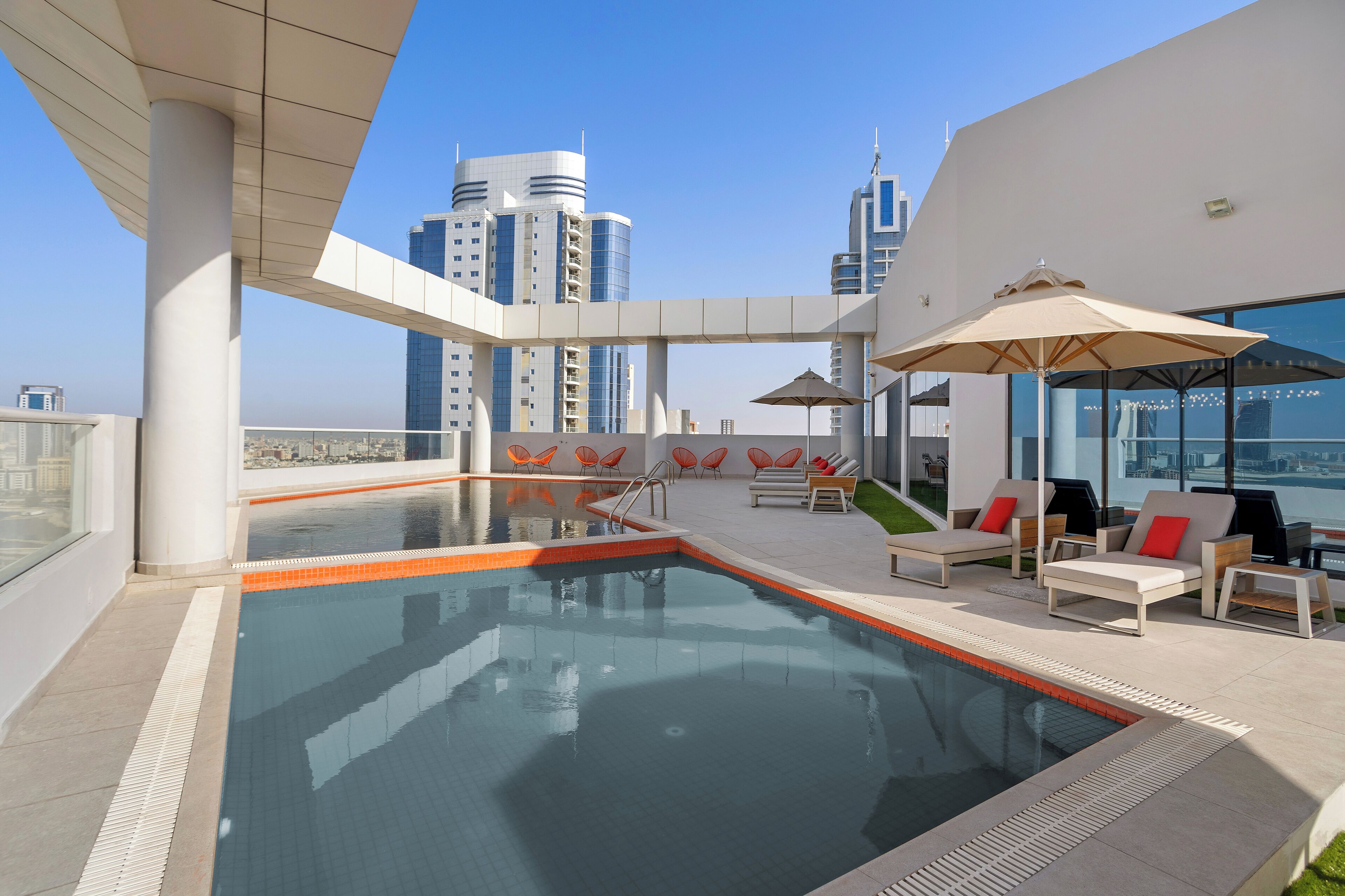 Una piscina en la azotea con barandilla de cristal, sillas anaranjadas y zona de lounge con sombrillas.