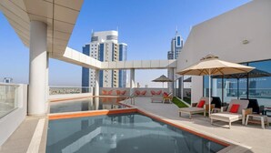 Un espace piscine aménagé sur le toit, avec des chaises longues et des parasols, entouré d’immeubles modernes.