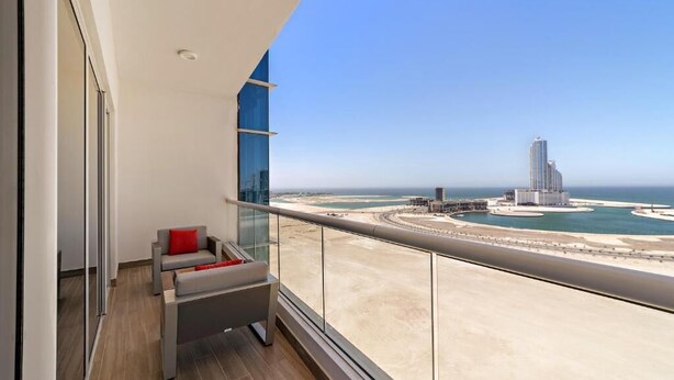 Un balcon offrant une vue sur un paysage urbain côtier, avec notamment un immeuble de grande hauteur et une marina circulaire.