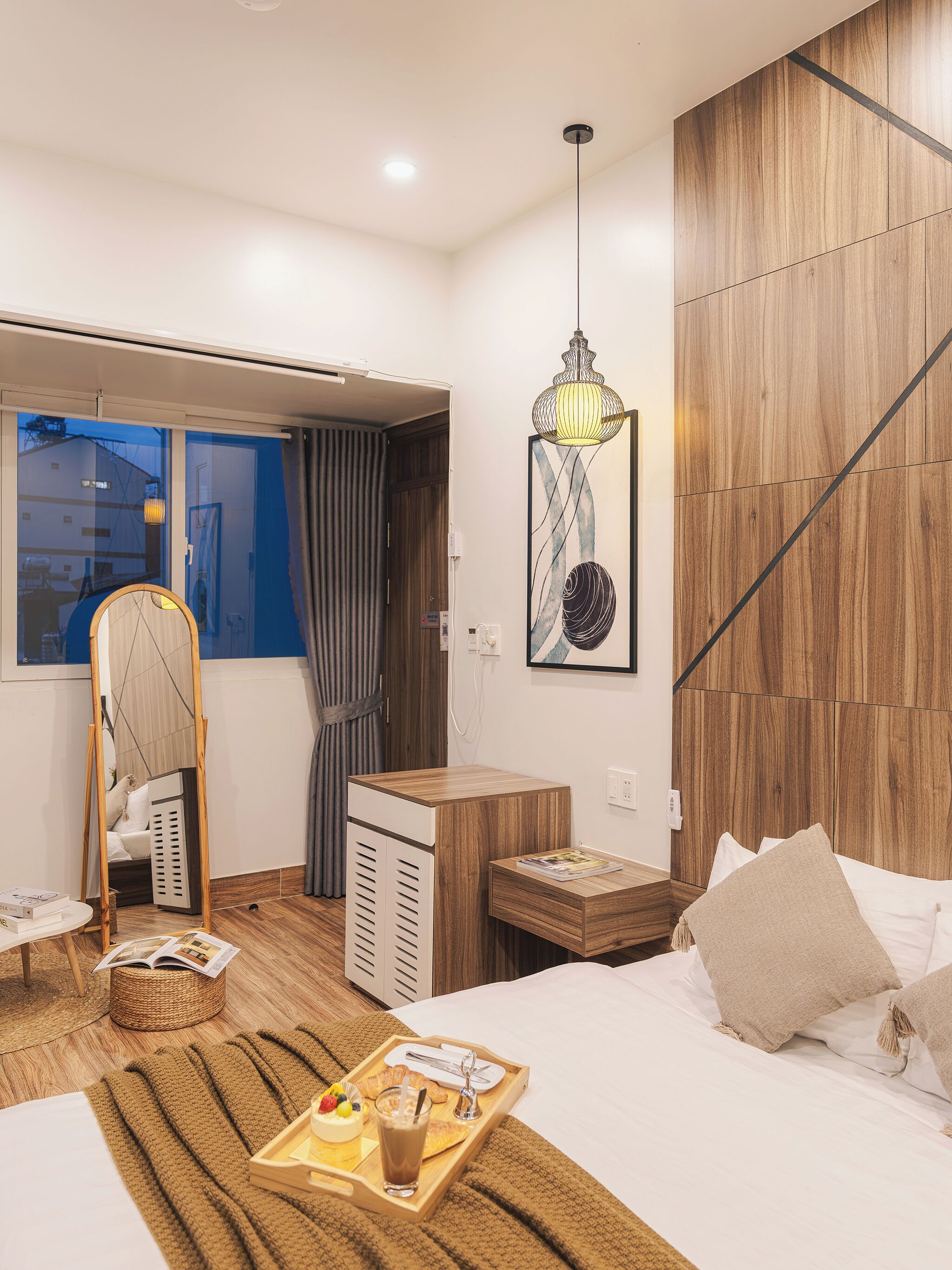 Ett modernt hotellrum med en säng, ett nattduksbord och ett litet skrivbord. Det finns ett fönster med gardiner, en hängande lampa och ett konstverk som hänger på väggen.