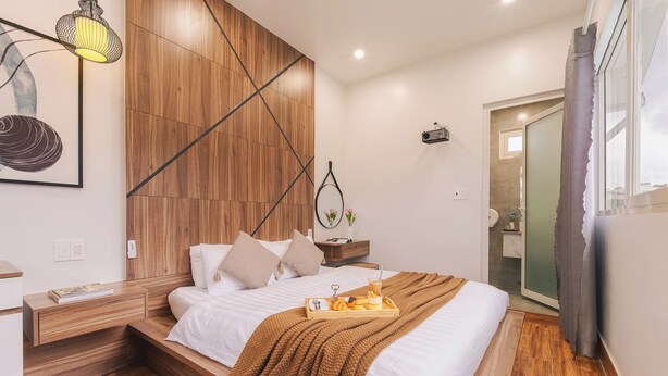A modern bedroom with a wooden headboard, a bedside table with a lamp, a tray of breakfast items, and a view of the bathroom through a glass door. - The Morris Central Hotel - O'TEL (Xuan Huong - Da Lat)