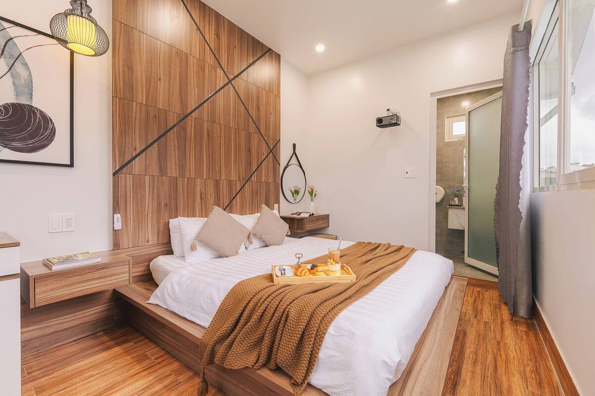 Une chambre moderne avec une tête de lit en bois, une table de chevet avec une lampe, un plateau contenant des articles pour le petit-déjeuner et une vue sur la salle de bain à travers une porte en verre.