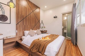 A modern bedroom with a wooden headboard, a bedside table with a lamp, a tray of breakfast items, and a view of the bathroom through a glass door. - The Morris Central Hotel - O'TEL (Xuan Huong - Da Lat)