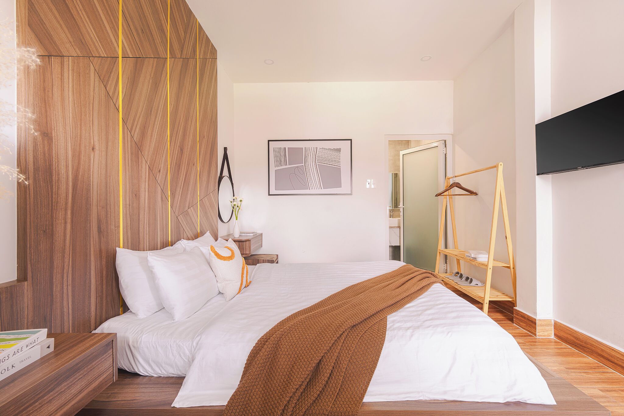 A modern bedroom with a wooden headboard, a large bed with white bedding, a brown throw blanket, a bedside table with a book, a wall-mounted TV, and a wooden shoe rack.