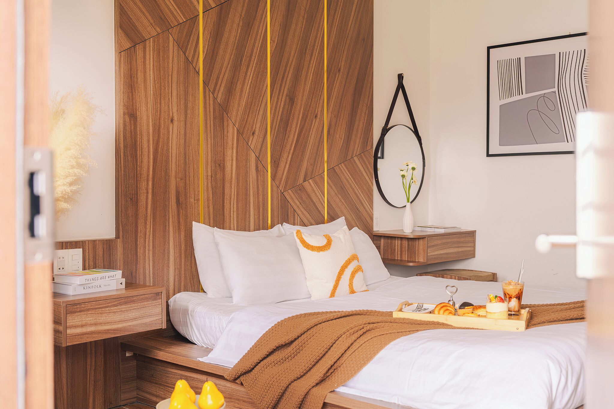 Quarto moderno com cabeceira de madeira, mesa de cabeceira com livros, luminária pendente e bandeja com comida e bebidas sobre a cama.