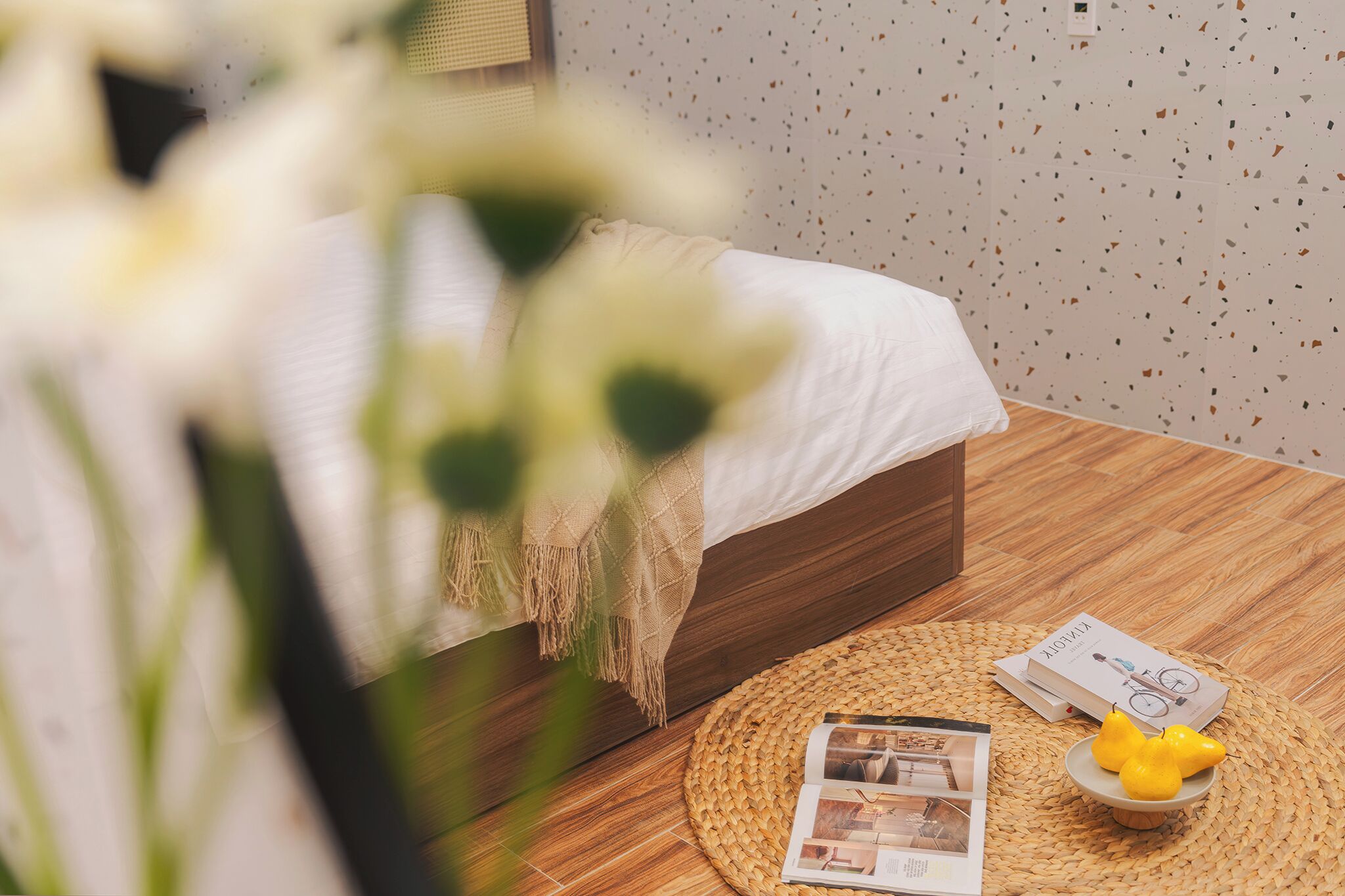 A neatly arranged bedroom with a bed, a woven round rug, a small table with a book and a yellow object, and a wall with a speckled pattern.