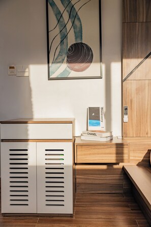 A modern hotel room with a wooden floor, a white cabinet with ventilation slits, a bedside table with a remote control, and a framed abstract painting on the wall. - The Morris Hotel - Central Dalat (Xuan Huong - Da Lat)