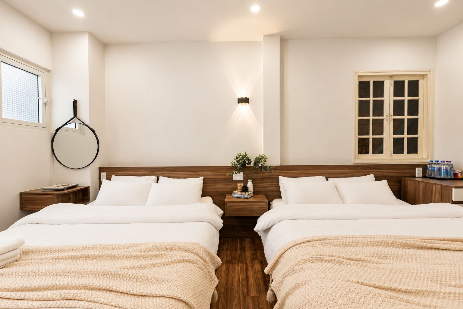 A hotel room with two beds, a bedside table with a lamp, a mirror, a window with blinds, and a wooden floor.