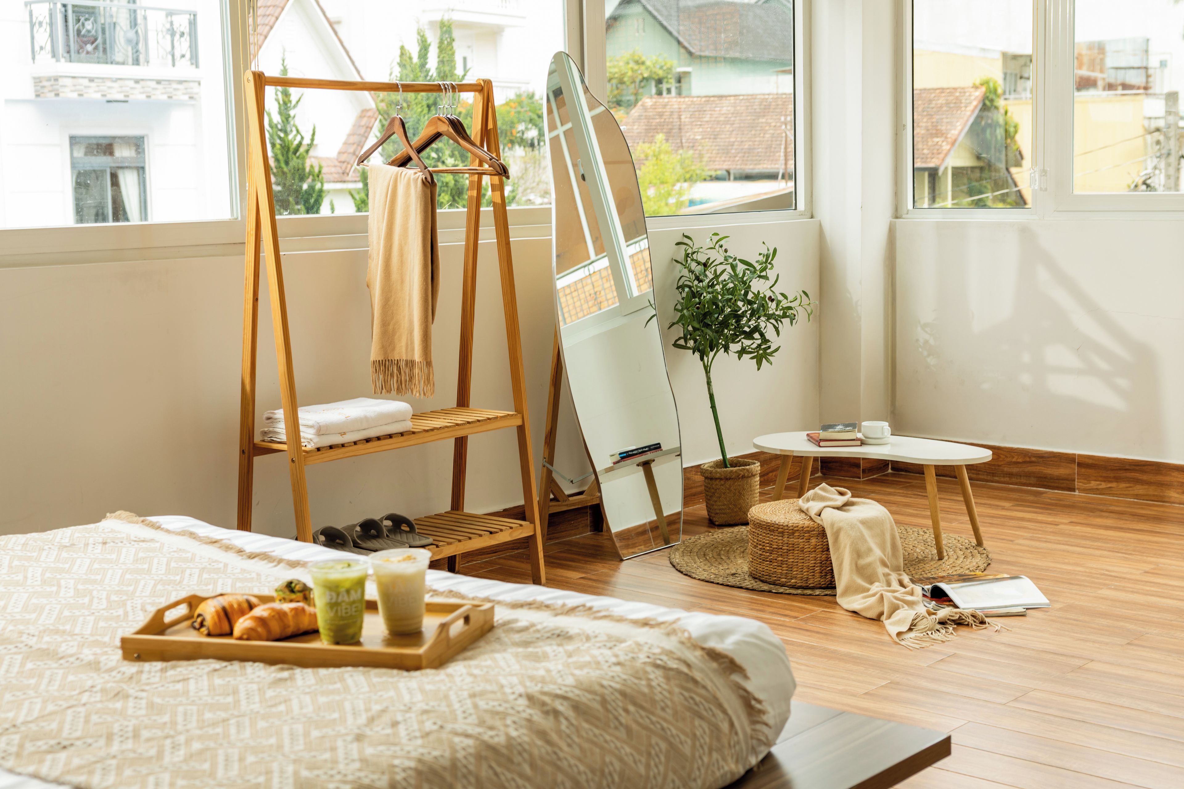 A room with a bed, a wooden drying rack, a mirror, a small table with a plant, and a window with a view of buildings.