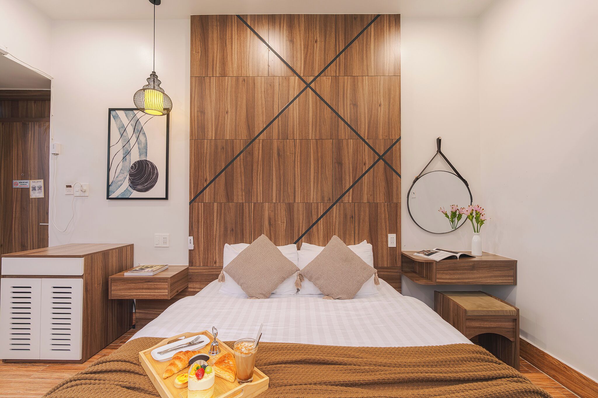 A modern hotel room with a wooden accent wall, a bed with white linens, a wooden nightstand, a small table with breakfast items, and a hanging light fixture.