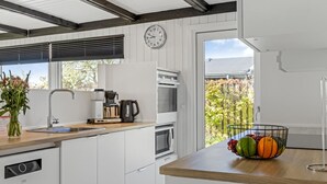 Une cuisine moderne avec des meubles blancs, un four encastré, une machine à café et un panier de fruits posé sur le plan de travail.