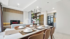 A modern dining area with a long table set for four, a television displaying 'NETFLIX', and a kitchen visible in the background. - Omira by Kozystay  - Cengkareng (Jakarta)