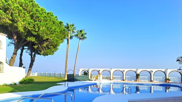 Ein Swimmingpool mit weiß gefliestem Deck, Palmen und ein Gebäude mit Bögen im Hintergrund.