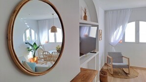 Ein Wohnzimmer mit großem Spiegel, Fernseher, Essbereich inklusive Stühle und Tisch sowie ein Fenster mit Vorhängen.