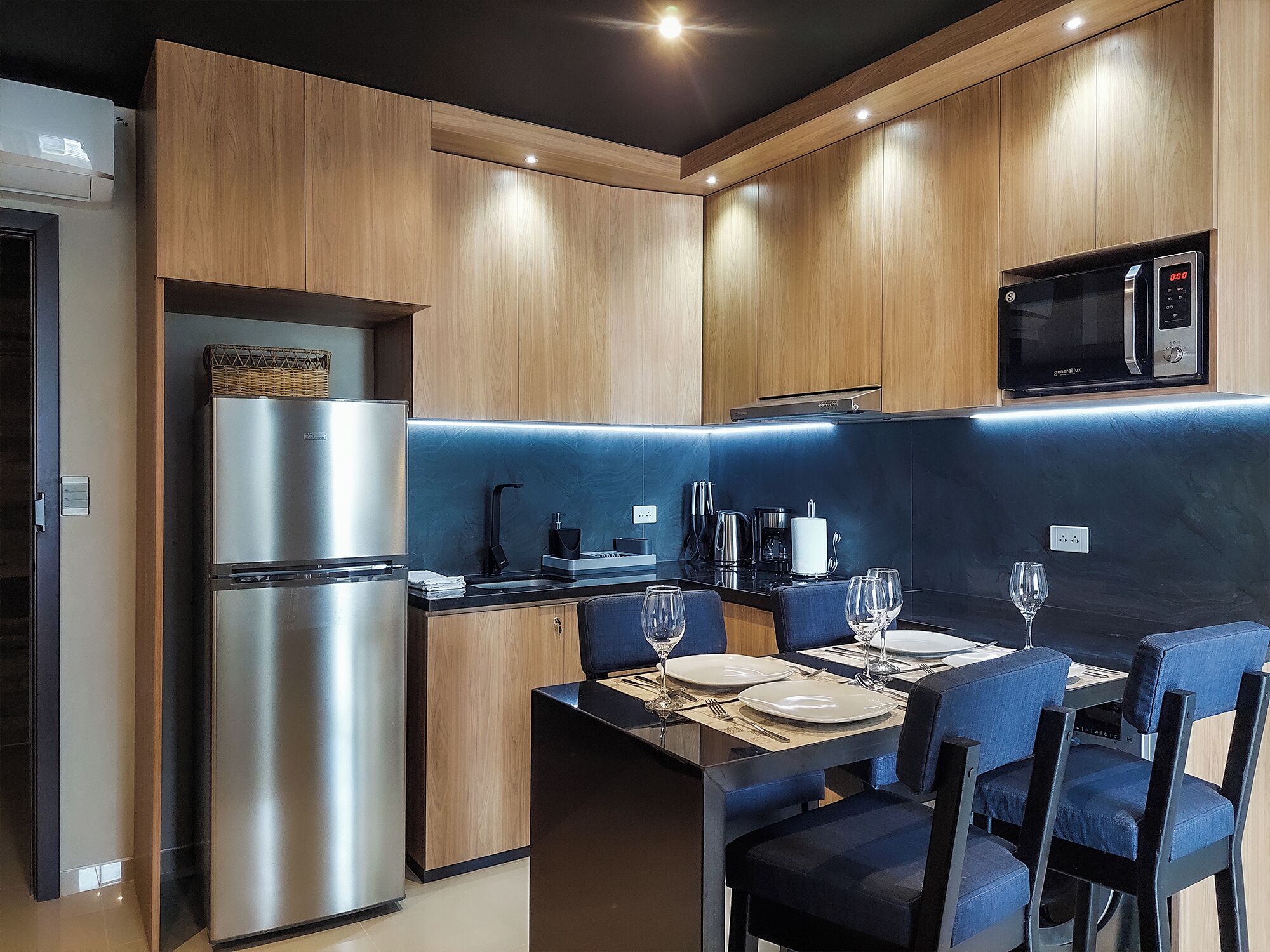 Una cocina moderna con armarios de madera, un refrigerador de acero inoxidable, un microondas y un comedor con sillas azules y una mesa para dos.