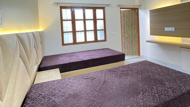 A room with a ceiling fan, a window with wooden frames, and a bed with a patterned mattress. - Host varanasi (Varanasi)