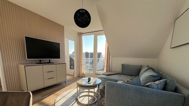 A modern living room with a flat-screen TV, a grey sofa, a round coffee table, and a large window offering a view of buildings.