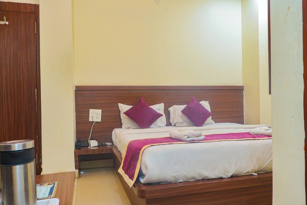 A hotel room with a wooden bed, white bedding, and purple pillows. There is a nightstand with a phone and a trash can.